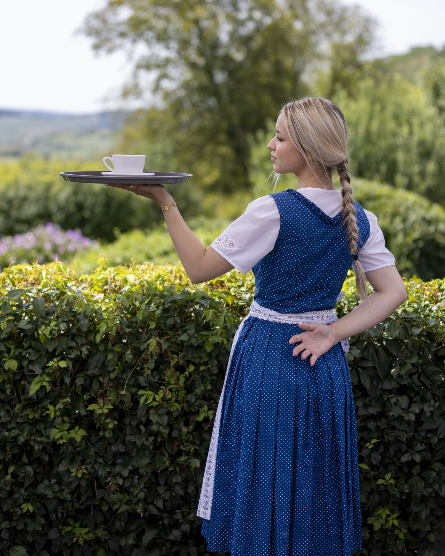 Kellnerin im blauen Dirndl serviert Kaffee vor grüner Hügellandschaft